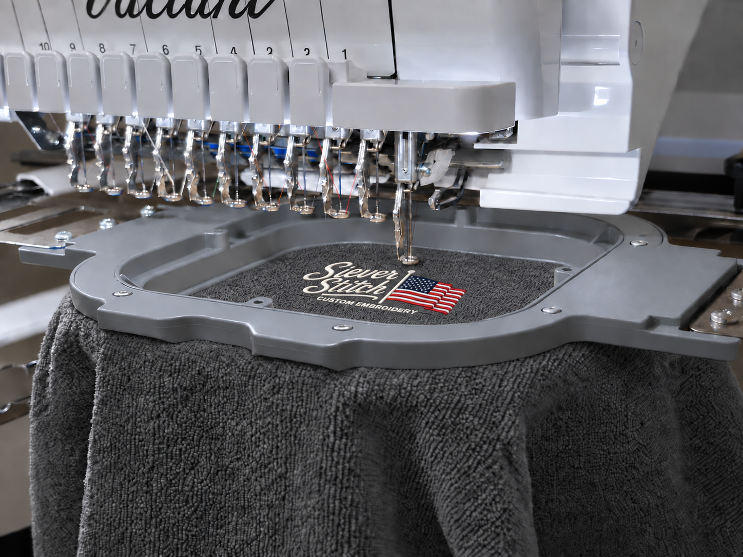 Edited photo of an embroidery machine stitching the Siever Stitch logo on a charcoal towel in a gray hoop